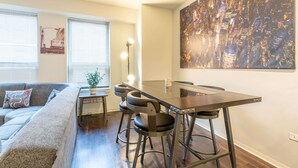 A modern living room with a sofa, a dining table with bar stools, a cityscape painting, and a lamp.
