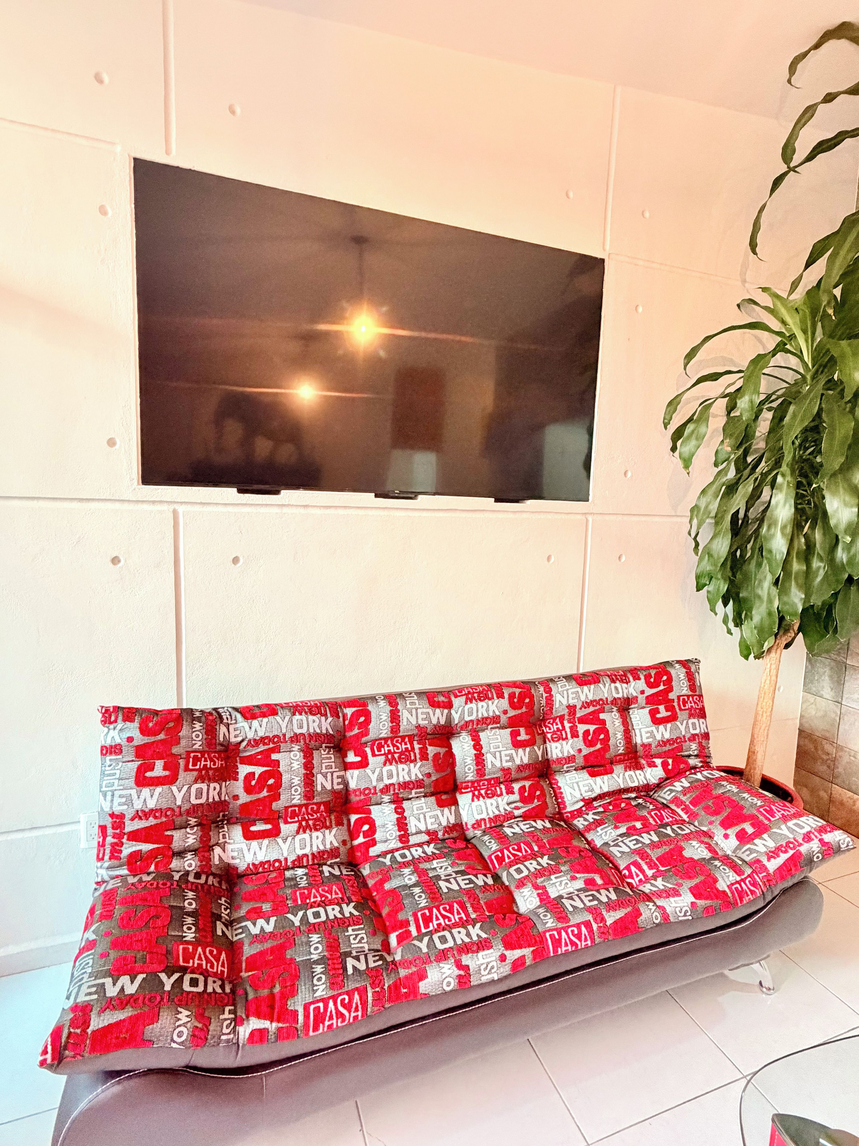 A sofa with a red and white patterned cushion, a window with a view of a building, and a potted plant to the right.