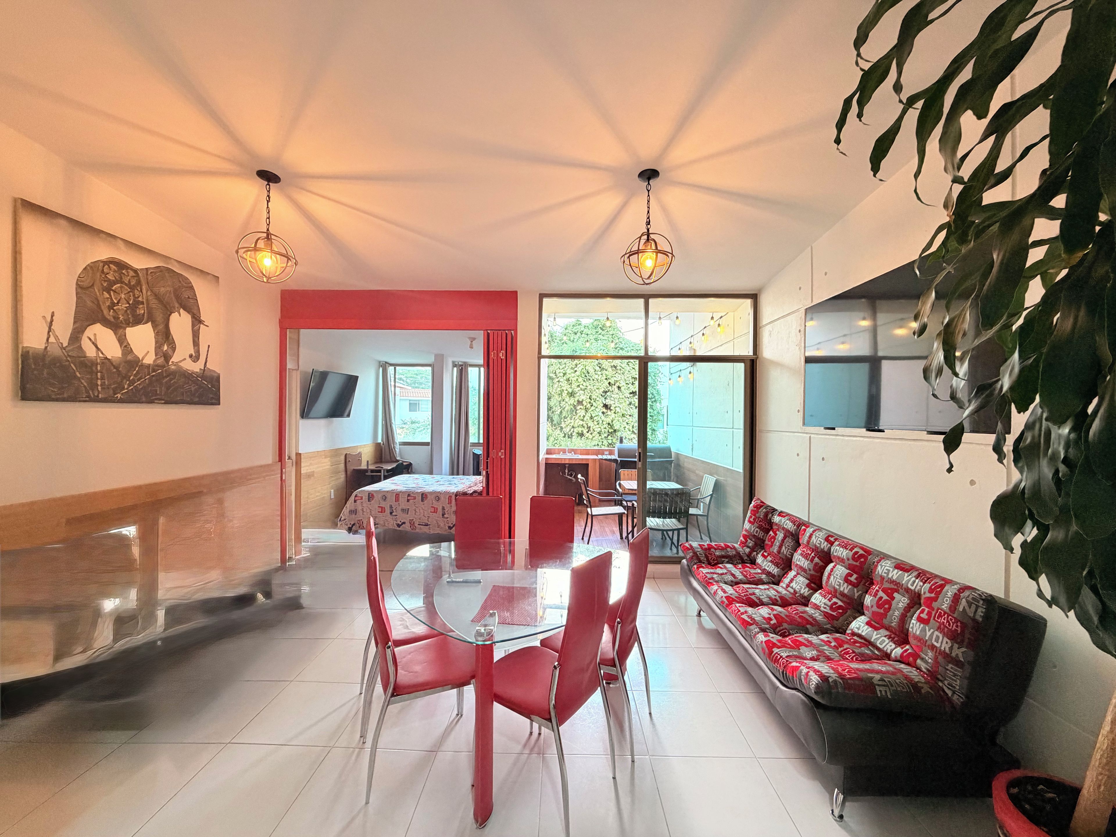 A dining area with a glass table and red chairs, a sofa with patterned cushions, and a television on the wall.