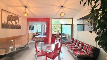 A dining area with a glass table and red chairs, a sofa with patterned cushions, and a television on the wall.