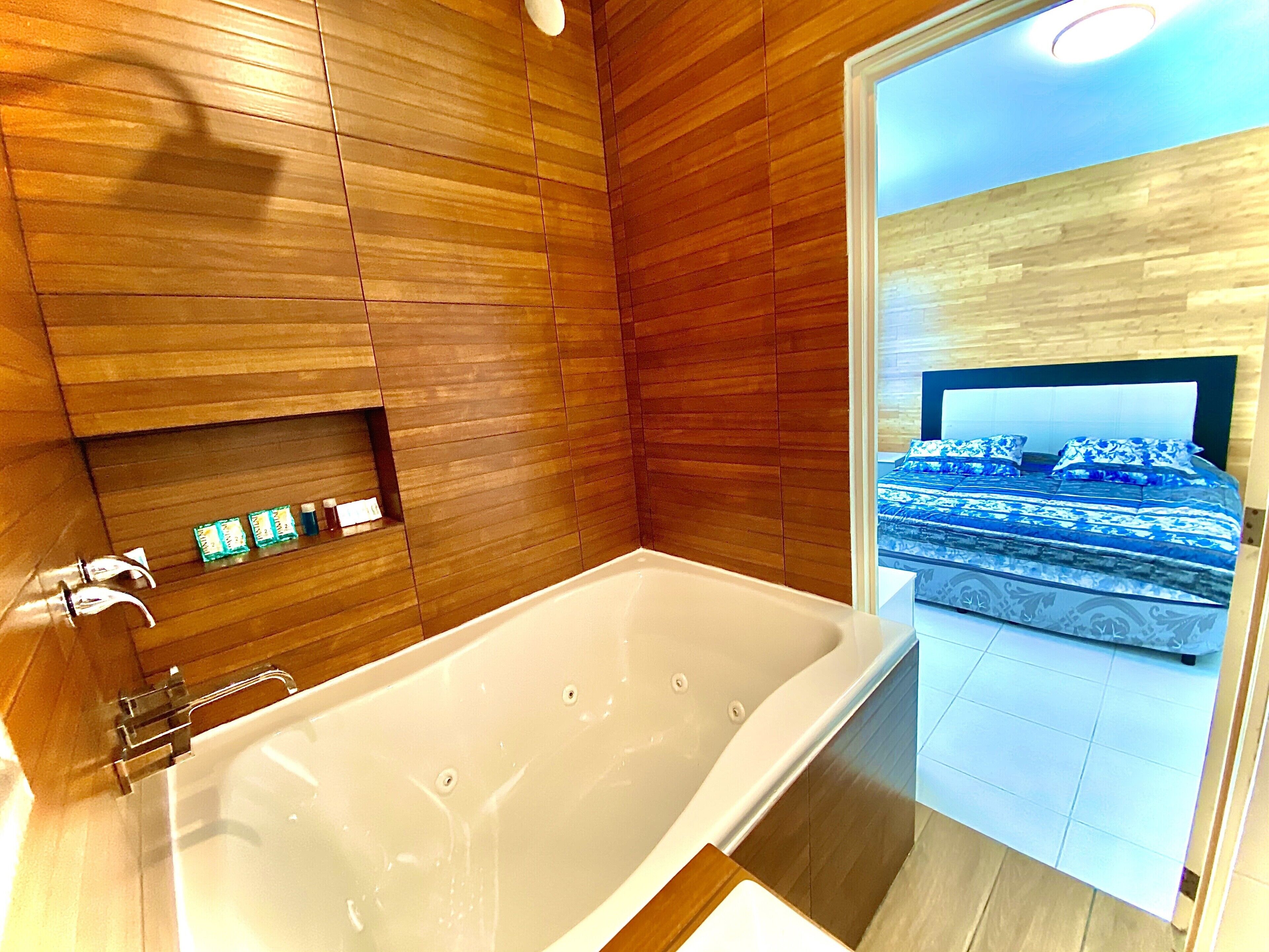 A modern bathroom with a large bathtub, wooden paneling, and a view of a bedroom with a bed and patterned bedding.
