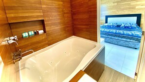A modern bathroom with a large bathtub, wooden paneling, and a view of a bedroom with a bed and patterned bedding. - Loft con Alberca Climatizada/2 Exclusivos Jacuzzis Premium/Terraza/Grill/Wi-Fi (Lomas Tetela)