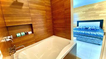 A modern bathroom with a large bathtub, wooden paneling, and a view of a bedroom with a bed and patterned bedding.