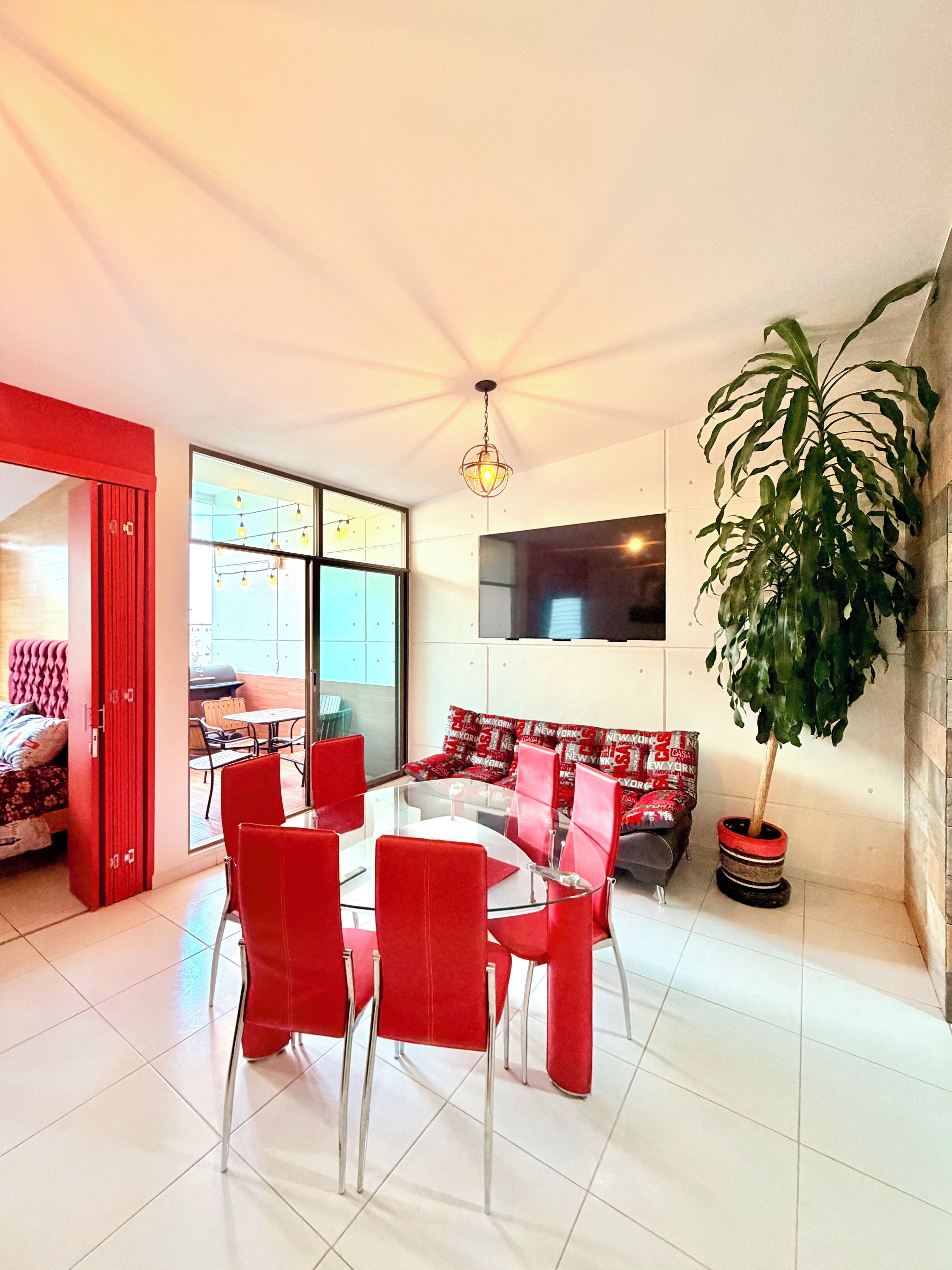 A modern living room with a glass dining table surrounded by red chairs, a large potted plant, and a red-framed sofa.
