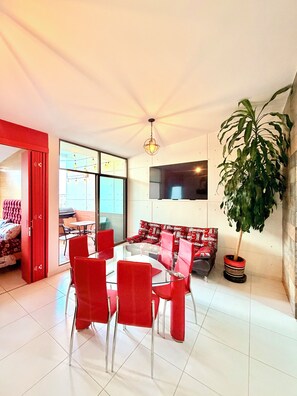 A modern living room with a glass dining table surrounded by red chairs, a large potted plant, and a red-framed sofa.