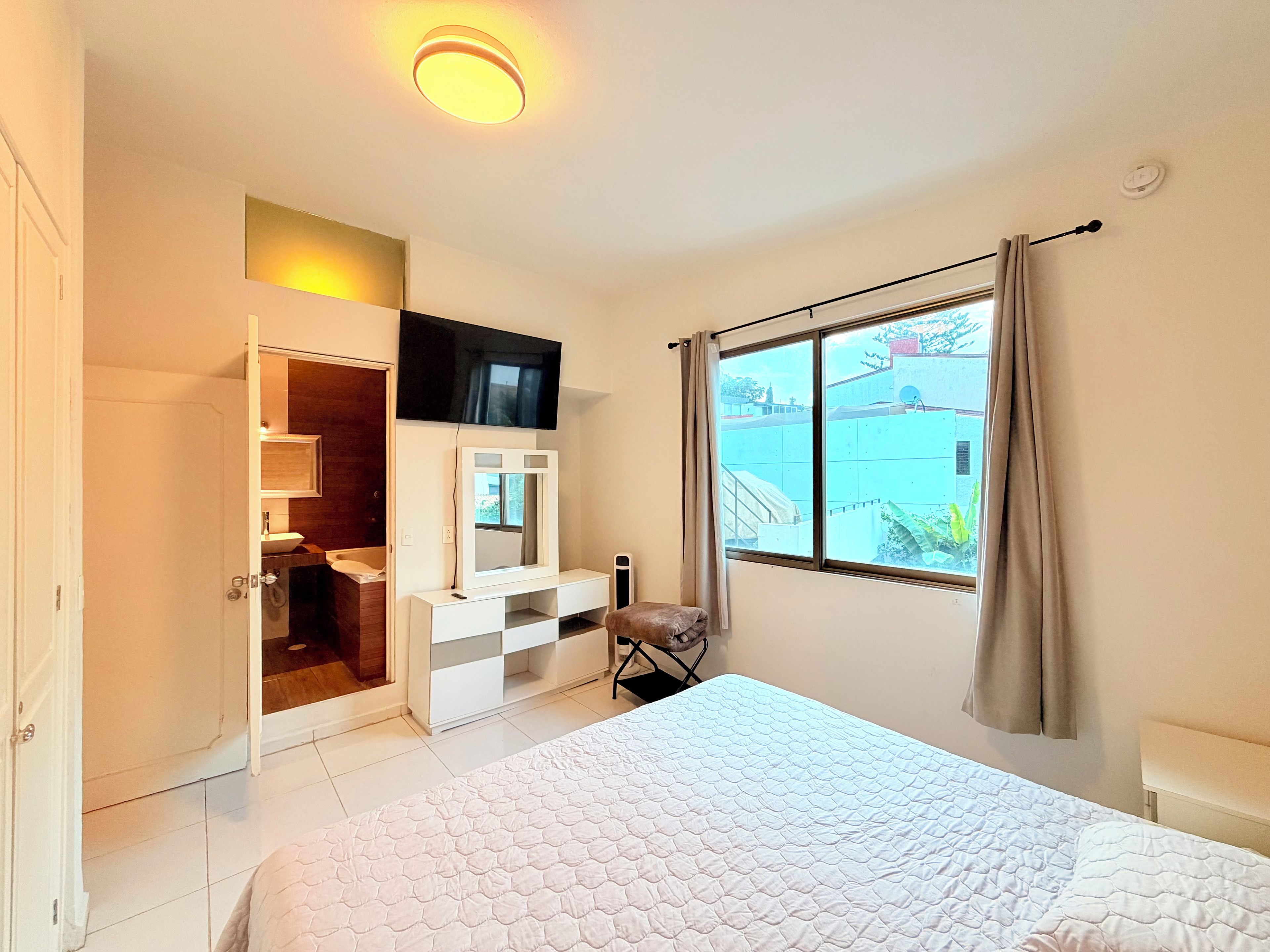Une chambre d’hôtel comprenant un lit, une coiffeuse avec un miroir, un téléviseur fixé au mur, une fenêtre avec des rideaux et une porte donnant sur une salle de bain.