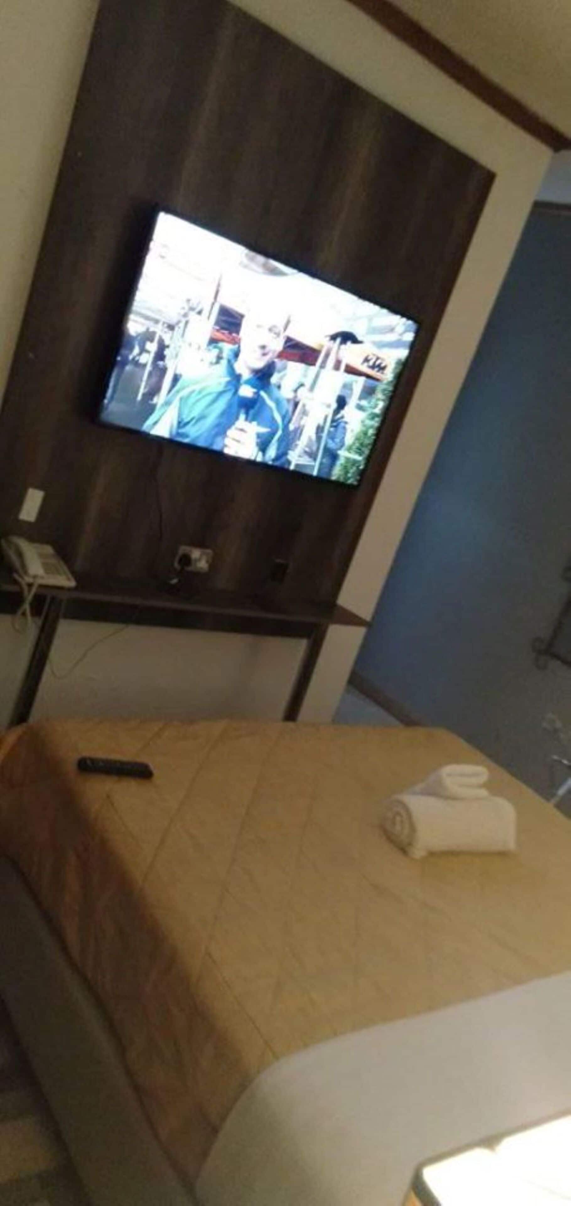 A hotel room with a bed, a TV mounted on the wall, a remote control on the bed, and a rolled towel on the bed.