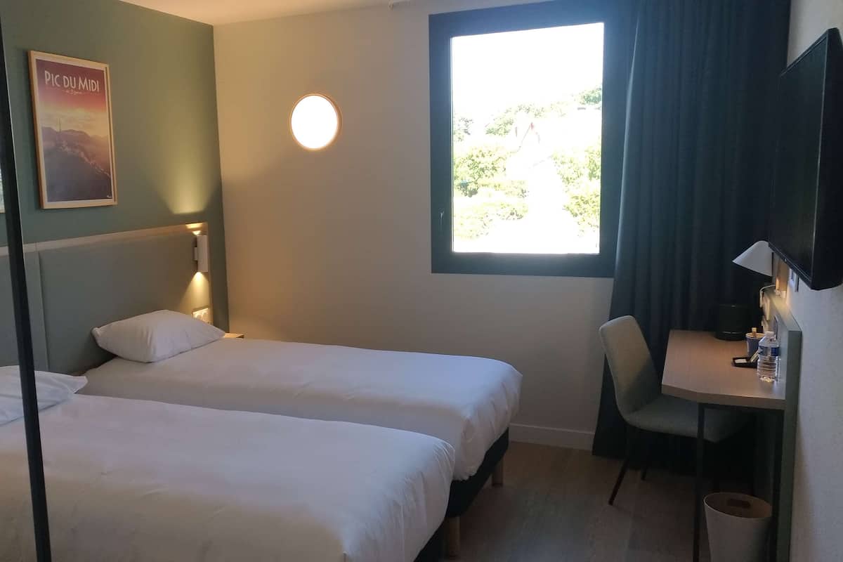 Une chambre d’hôtel avec deux lits, un bureau, une chaise, une télévision et une fenêtre donnant sur un paysage verdoyant.