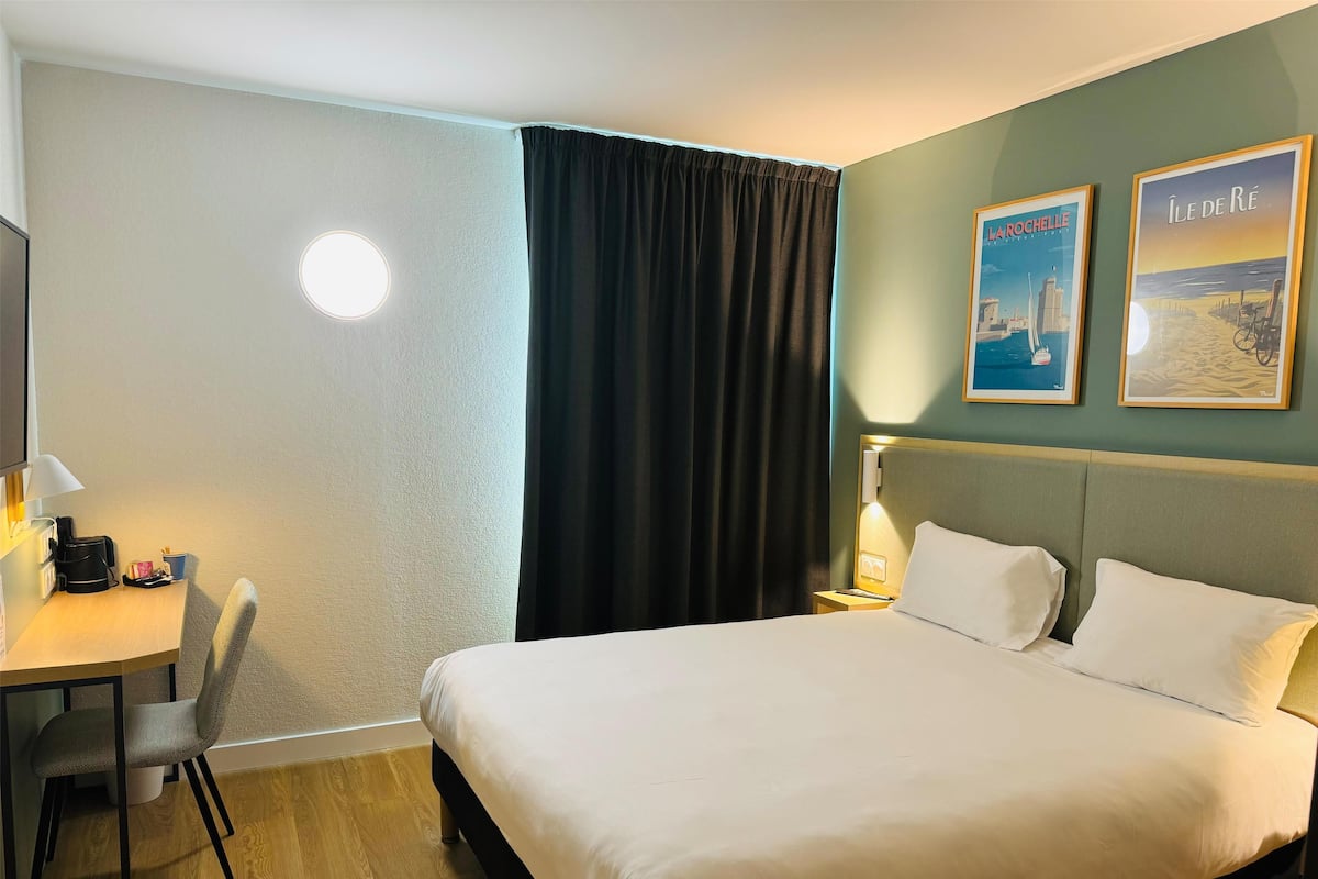 Une chambre d’hôtel avec un lit, un bureau et une chaise, une lampe et deux tableaux encadrés au mur.