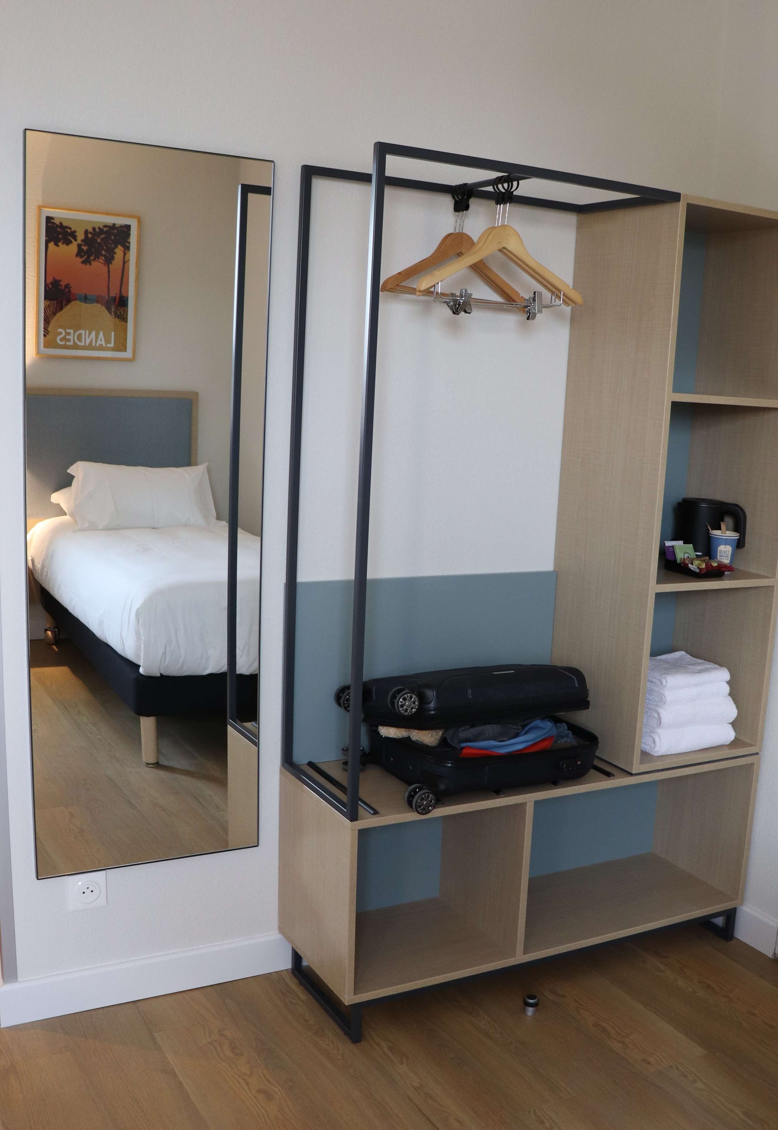 A room with a bed, a wardrobe, a mirror, and a shelf with a suitcase and folded clothes.