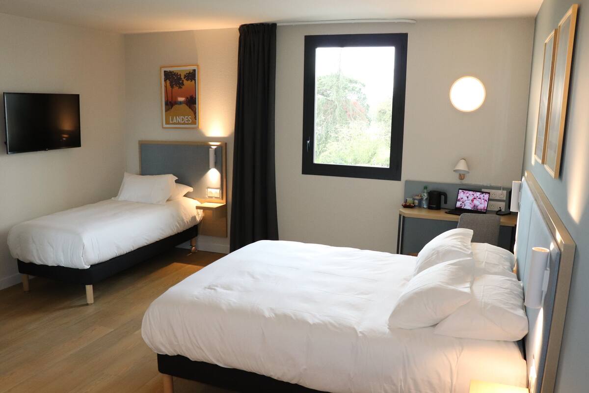 Une chambre d’hôtel avec deux lits simples, un bureau avec un ordinateur, une télévision et une fenêtre avec des rideaux.
