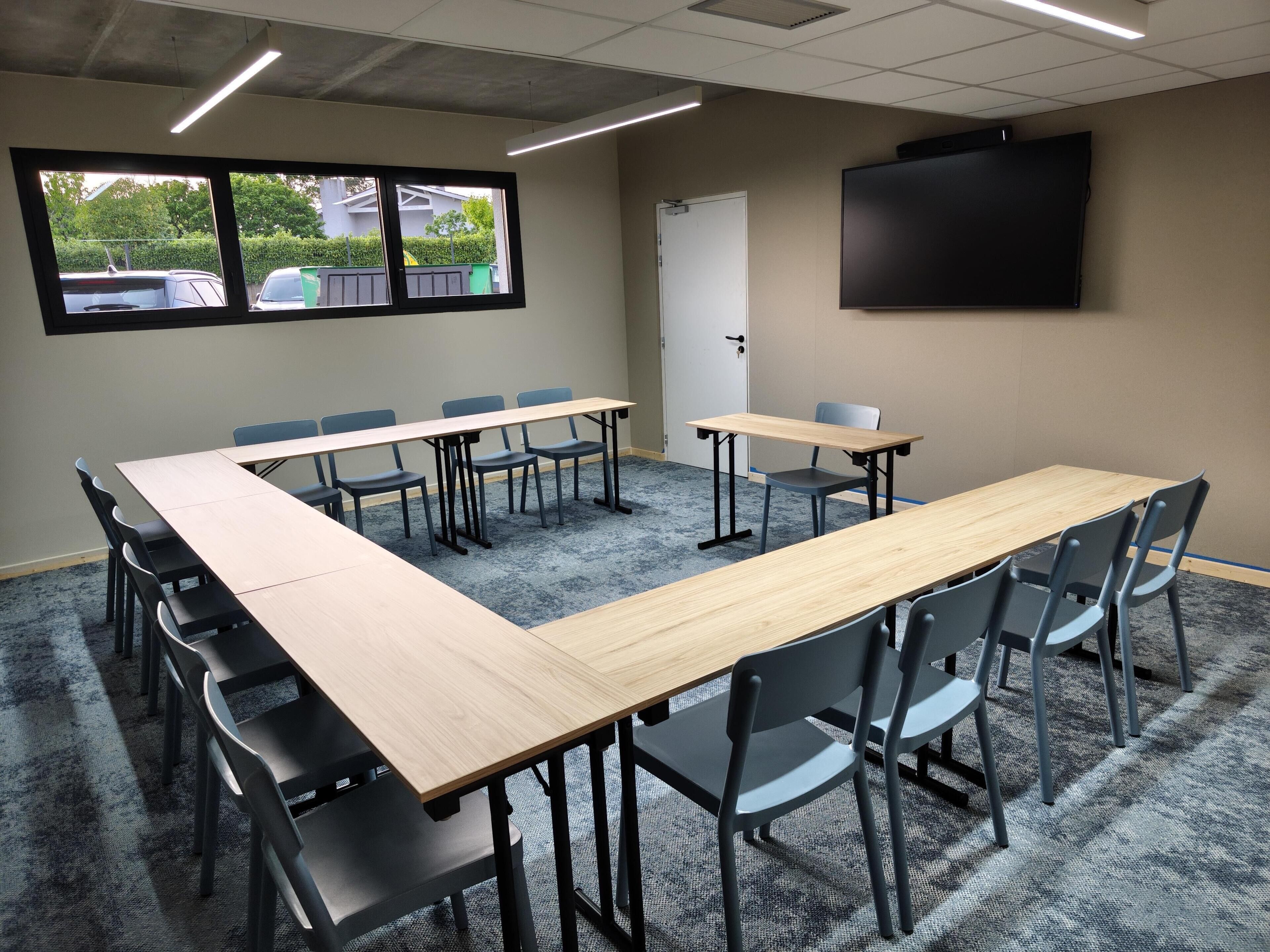 A conference room with a long table, chairs, a projector screen, and a window with a view of parked cars and greenery.
