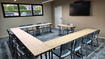 A conference room with a long table, chairs, a projector screen, and a window with a view of parked cars and greenery.