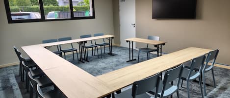 Un salón de conferencias con una larga mesa, sillas, una pantalla de proyector y un ventanal con vistas a coches aparcados y zonas verdes.
