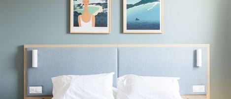 Habitación de hotel con cama, almohadas blancas, cabecera de madera, dos cuadros enmarcados en la pared y mesitas de noche con un control remoto y un enchufe.