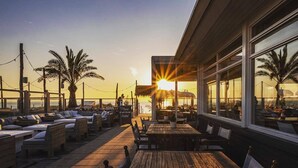 An outdoor seating area with wooden tables and chairs, palm trees, and a view of the sunset over the ocean. - Bungalow on vacation park near the beach Noordwijk (Noordwijkerhout)