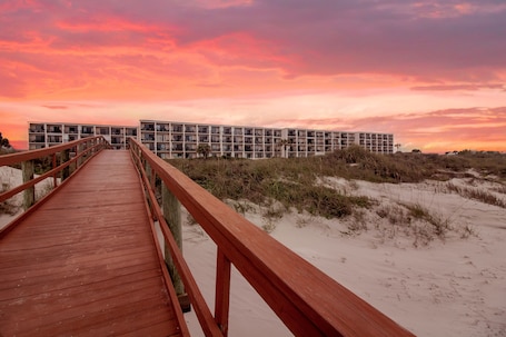 A wooden boardwalk leading to a modern building with multiple balconies, situated on a sandy beach with dunes in the background.