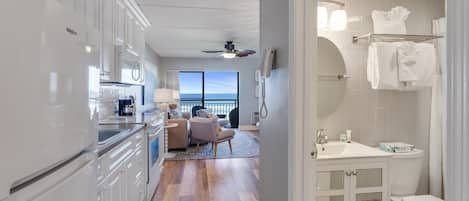 A modern kitchen with white cabinetry, a dining area with a gray sofa, and a bathroom with a white sink and toilet.