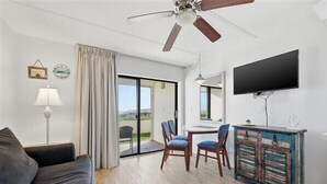 A modern living room with a sofa, a dining table with chairs, a ceiling fan, a TV mounted on the wall, and a sliding glass door leading to an outdoor area.