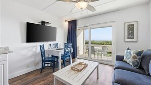 Un salon moderne doté d’un ventilateur de plafond, d’un téléviseur à écran plat fixé au mur, d’un coin repas avec des chaises bleues, d’un canapé agrémenté d’un coussin à motifs et d’une porte-fenêtre coulissante donnant sur une terrasse extérieure.