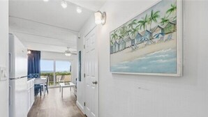 Un couloir donnant sur une vue de plage, un coin repas avec des chaises et une table, et une porte blanche.