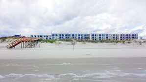 Een strandhotel met een pier die naar het gebouw leidt, een zandstrand en duinen.