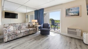 Une pièce comprenant un canapé, un fauteuil, une table avec une lampe, un téléviseur et une grande fenêtre donnant sur un beau paysage.
