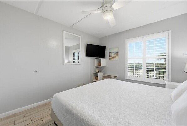 Ein Schlafzimmer mit einem Bett, einem Deckenventilator, einem Fernseher, einem Fenster mit Jalousien und einem kleinen Tisch mit einer Lampe.