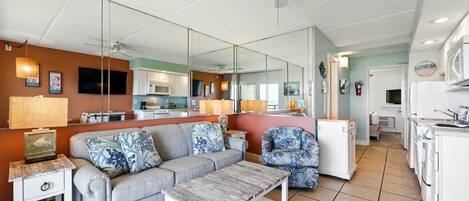 A modern living room with a grey sofa, a coffee table, and a mirrored wall reflecting the kitchen area.