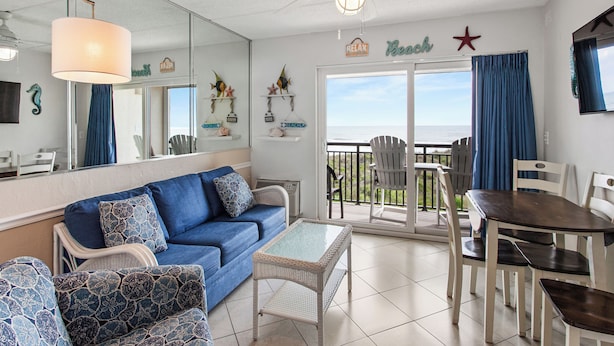 A living room with a blue sofa, white wicker furniture, a glass-top coffee table, and a dining table with chairs. There is a large mirror on the wall, a ceiling fan, and a view of the outdoors through a sliding glass door. - Beacher's Oceanfront Balcony Suite with onsite Front Desk and Staff (St. Augustine)