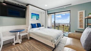 Un dormitorio con cama, una mesita, un sofá, un televisor y un balcón con vistas al cielo y las nubes.