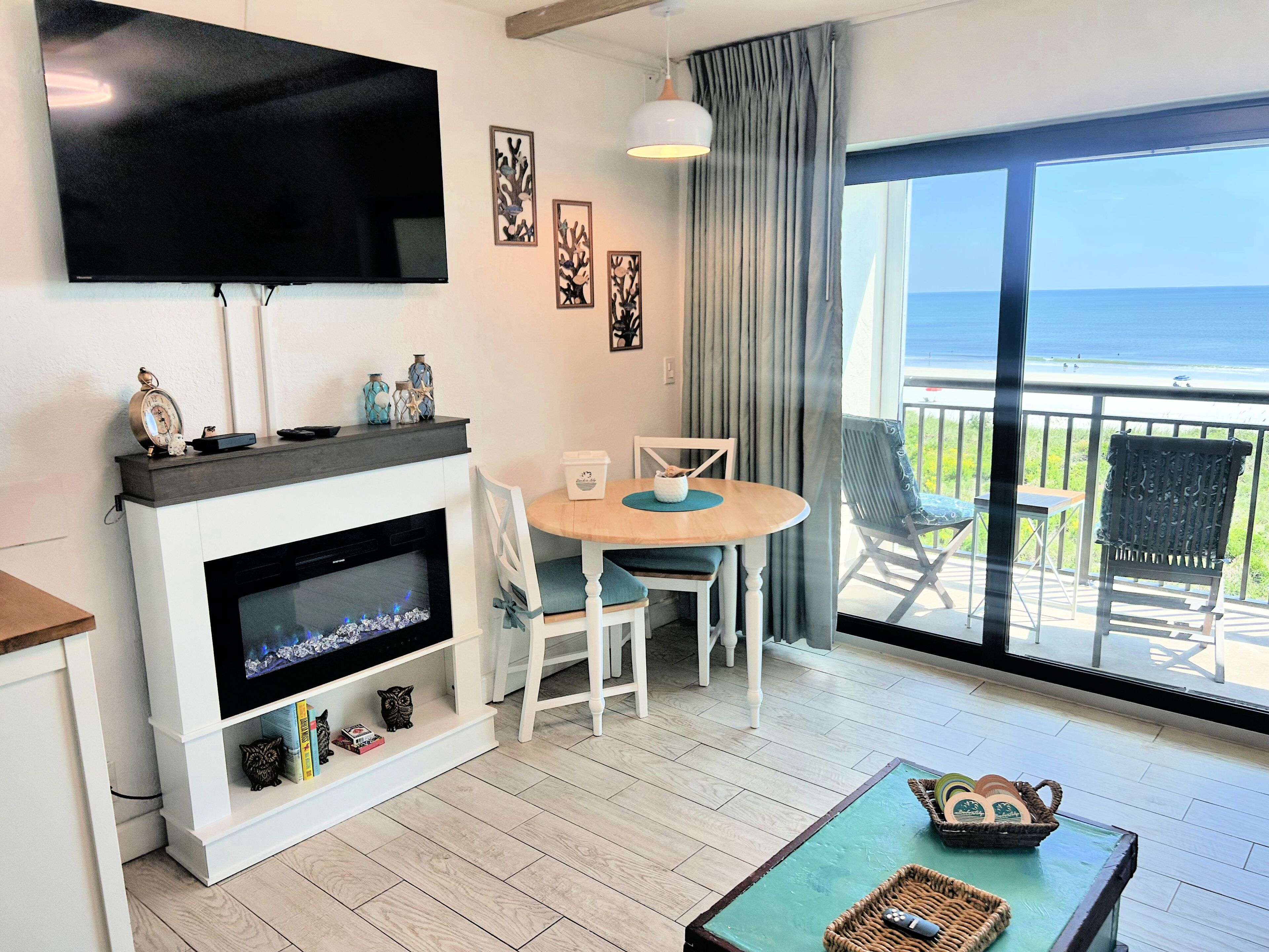 A living room with a fireplace, a television, a dining table with chairs, and a view of the ocean through large windows.