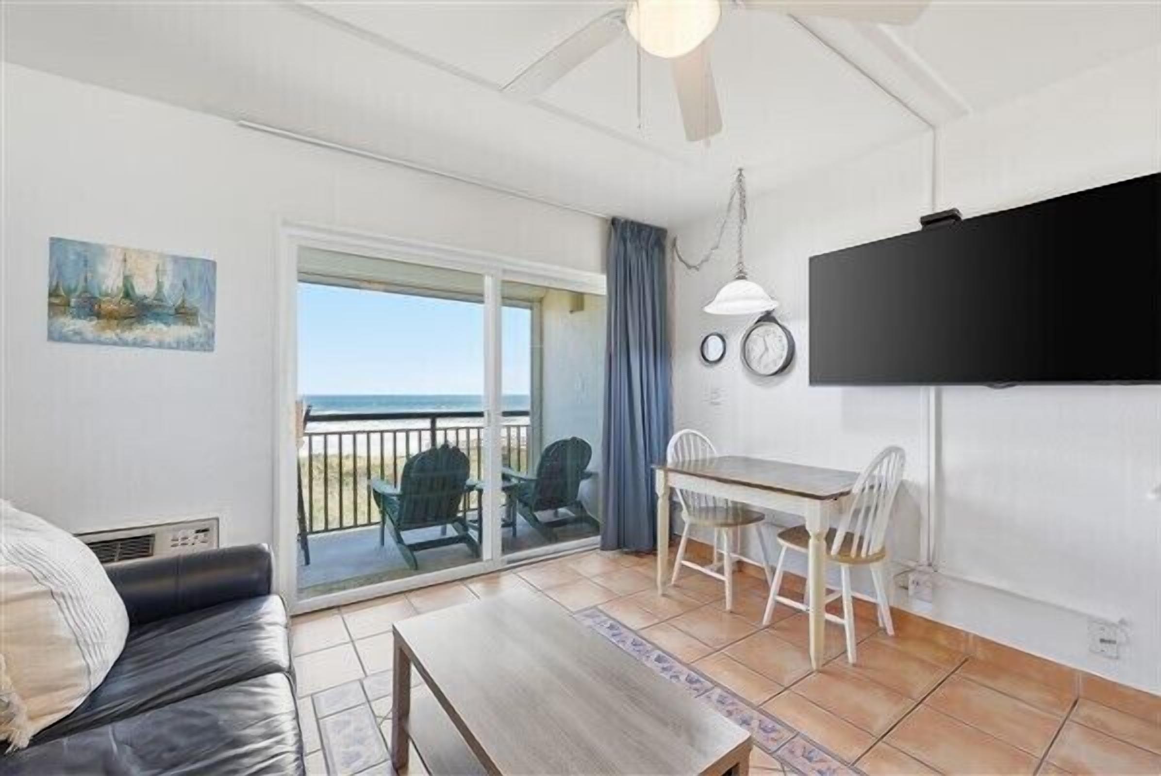 Une pièce comprenant un canapé, une table à manger avec des chaises, un téléviseur fixé au mur et une vue sur l’océan par une porte-fenêtre coulissante.