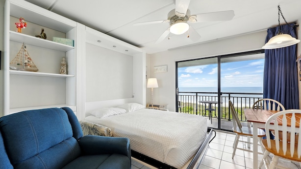 Een slaapkamer met een bed, een blauwe fauteuil, een houten tafel met stoelen, een plafondventilator en een balkon met uitzicht op de lucht en de zee.