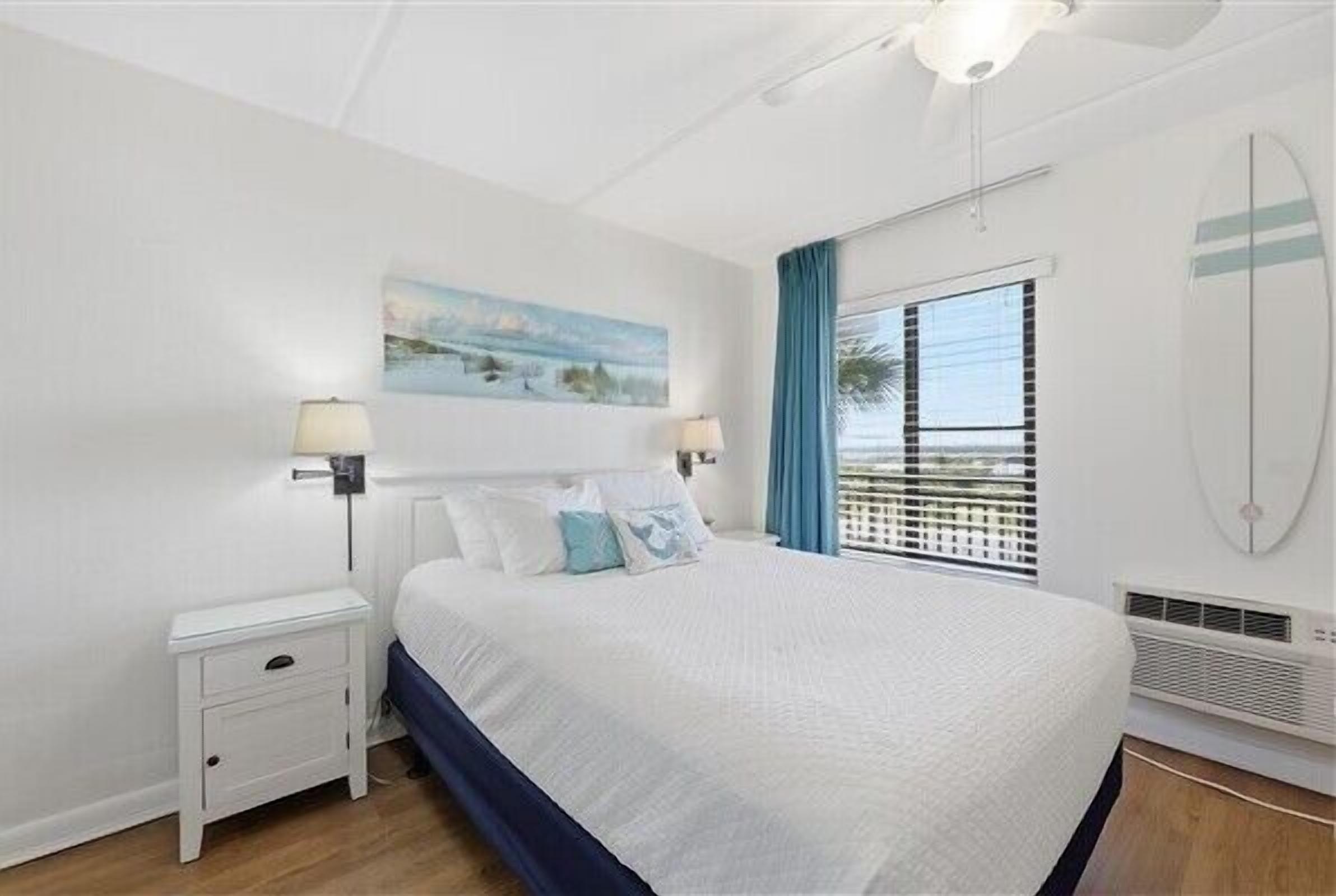 A bedroom with a bed, a nightstand, a wall-mounted lamp, a window with curtains, and a beach-themed painting.
