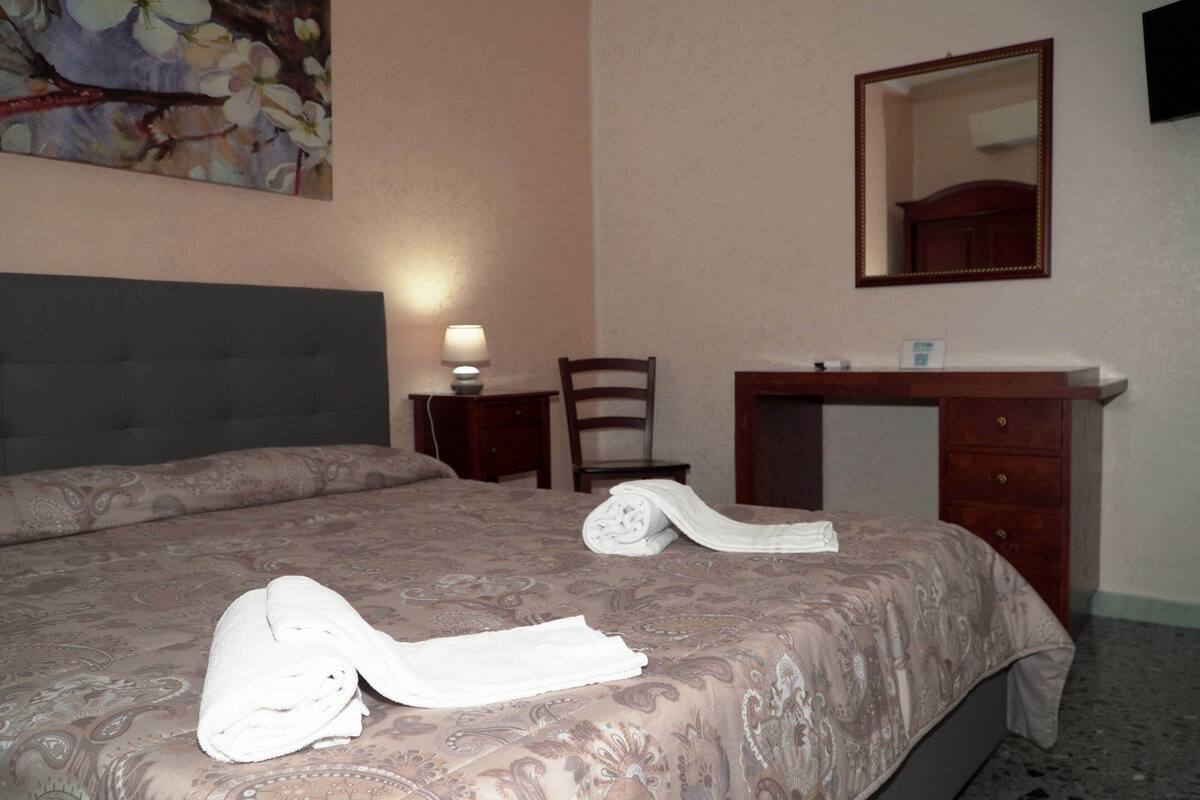 A hotel room with a bed, a desk, a chair, a mirror, and a painting on the wall.