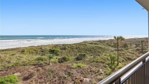 Vue sur la plage depuis un balcon doté d’une balustrade en bois, avec une plage de sable et l’océan en arrière-plan.