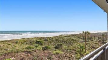 Vista a la playa desde un balcón con barandilla de madera, playa arenosa y océano al fondo.