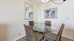 Un coin repas avec une table en verre entourée de chaises en osier, un luminaire suspendu au plafond et deux tableaux encadrés accrochés aux murs.