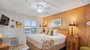 Een slaapkamer met een bed, een plafondventilator, een raam, een wandlamp en een klein tafeltje met een lamp.