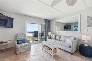 A living room with a sofa, armchair, coffee table, TV, and a balcony with a view.