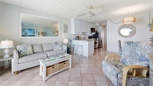 A living room with a sofa, coffee table, and wicker chairs. There is a kitchen area with cabinets and a ceiling fan. - Beacher's Oceanfront Balcony Suite with Onsite Front desk and staff (St. Augustine)