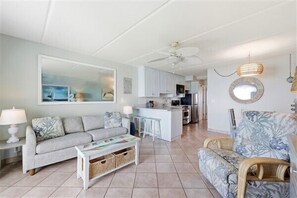 A living room with a sofa, coffee table, and wicker chairs. There is a kitchen area with cabinets and a ceiling fan. - Beacher's Oceanfront Balcony Suite with Onsite Front desk and staff (St. Augustine)