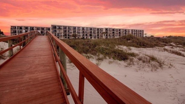 A wooden boardwalk leading to a modern building with multiple balconies, situated on a sandy beach with dunes in the background.