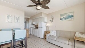 A spacious living area with a sofa, dining table, and chairs, a kitchen in the background, and a ceiling fan.