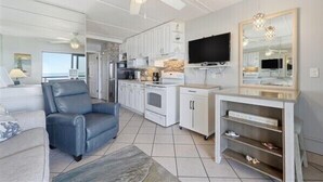 A modern living room with a blue leather chair, a TV, and a kitchen area with white cabinets and a stone backsplash.