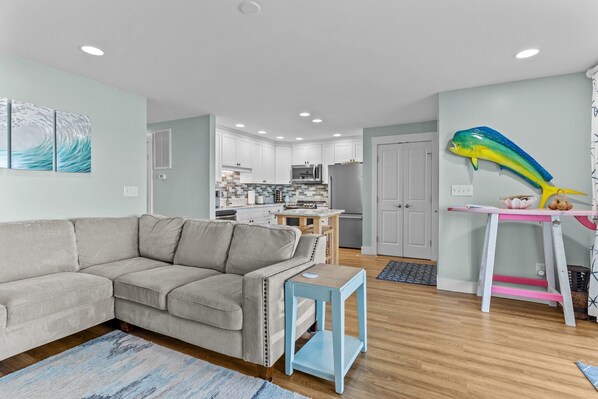 Un salón moderno con un sofá seccional gris, una mesita auxiliar azul y una cocina al fondo con armarios blancos y una gran decoración con forma de pez.