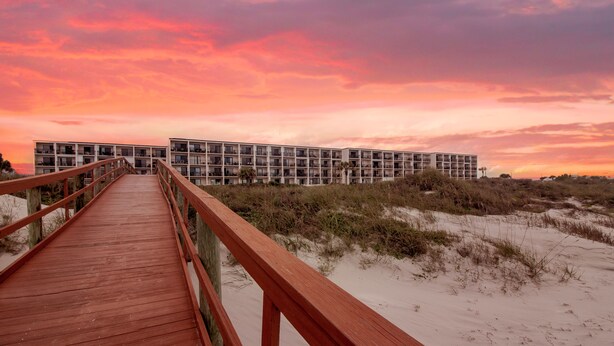 Une promenade en bois mène à un bâtiment moderne doté de nombreux balcons, situé sur une plage de sable avec des dunes en arrière-plan.