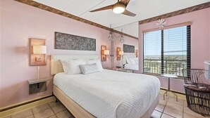 A bedroom with a large bed, bedside lamps, a ceiling fan, a window with blinds, and a small table with a chair. - Beacher's Oceanfront Balcony Suite with Onsite Front desk and staff (St. Augustine)