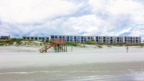 A beachfront hotel with a staircase leading to a deck, surrounded by sand dunes and a cloudy sky.